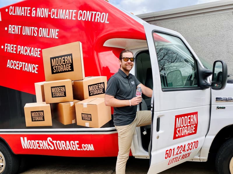 Modern Storage employee standing at open truck door with boxes stacked beside a branded red moving truck promoting climate control and online rentals