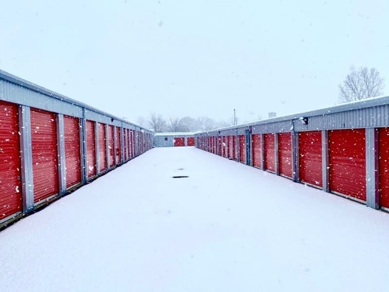 Snow-covered outdoor storage units at Modern Storage, showing red roll-up doors lining a wintery drive aisle, ideal for seasonal storage tips or winter weather preparation.