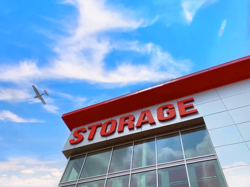 Upward view of a Modern Storage facility with red signage under a vibrant summer sky and an airplane flying above