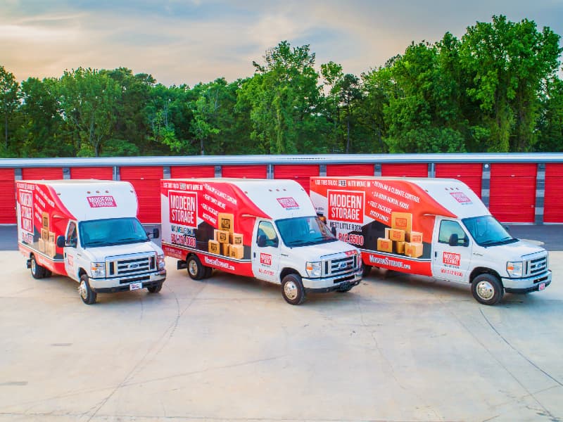 Three Modern Storage® branded moving trucks parked outside red roll-up storage units surrounded by trees