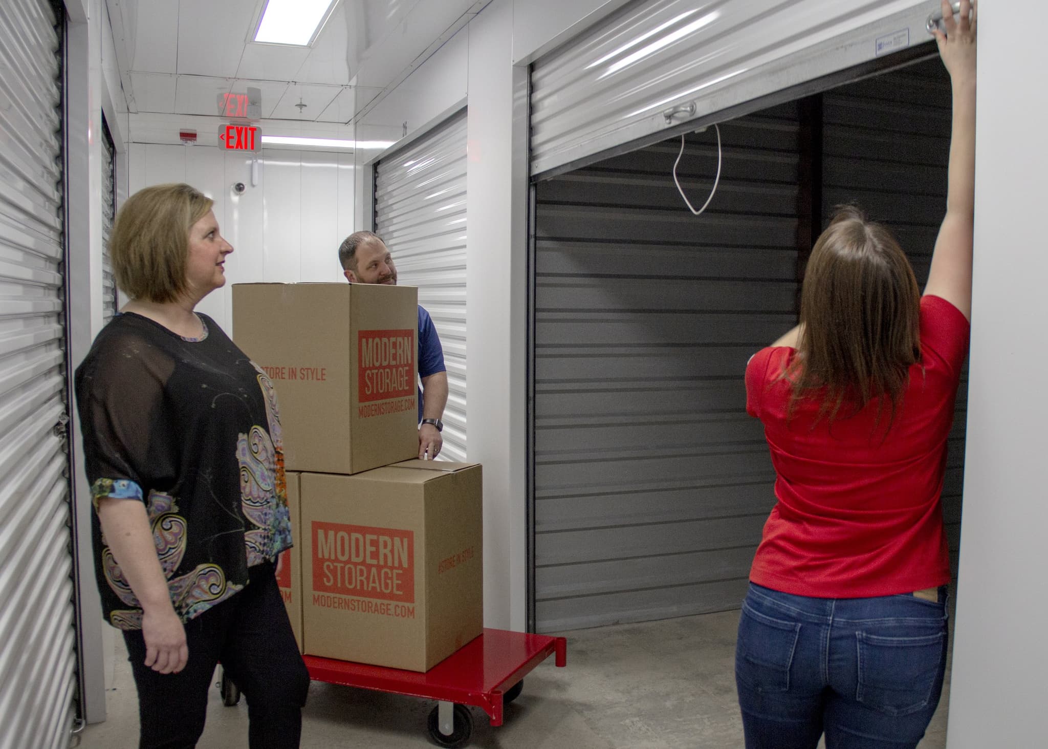 Three people using a Modern Storage® unit to load moving boxes and furniture during a downsizing transition, with a branded box cart and open roll-up door.