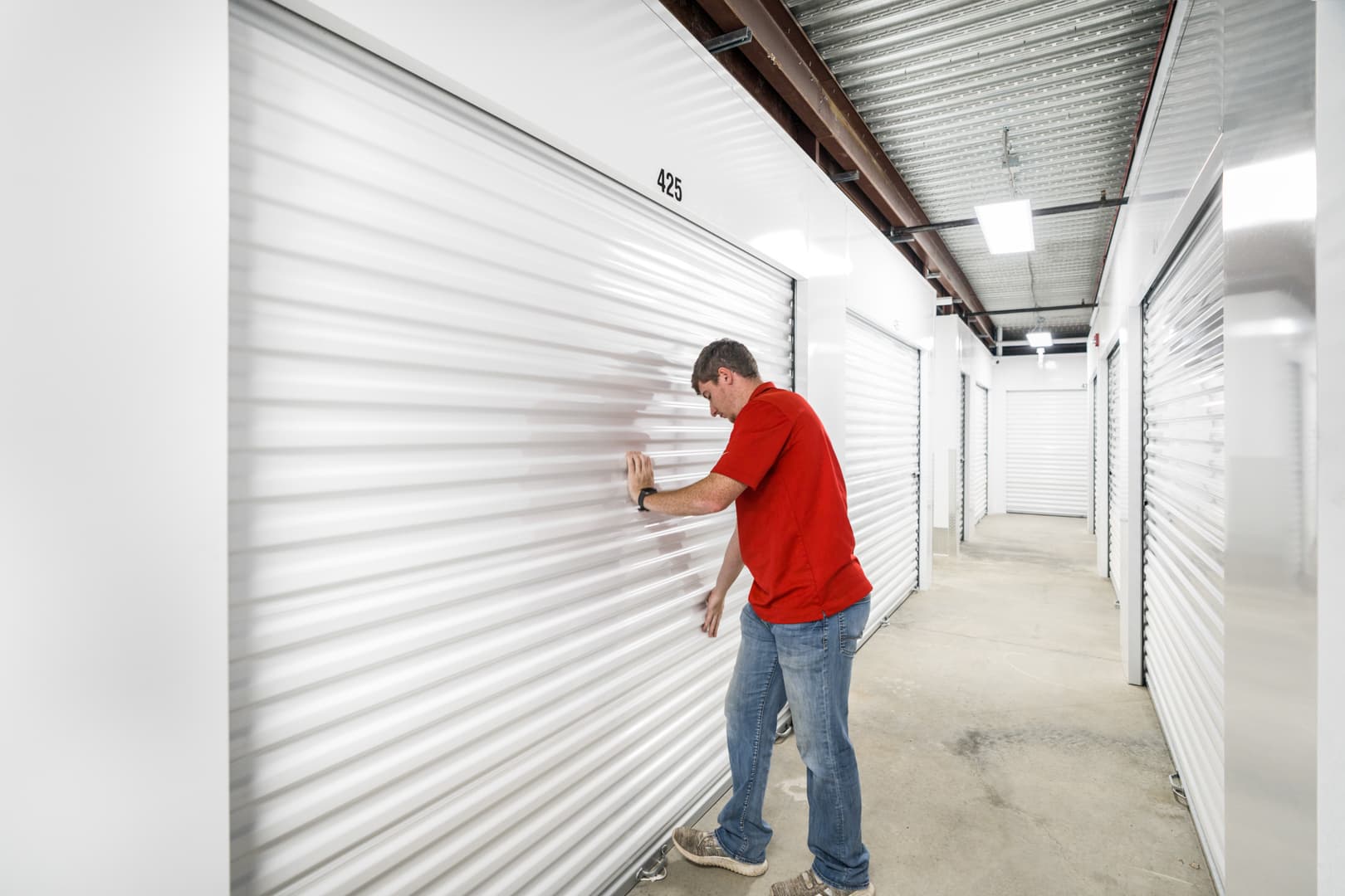 Man in a red shirt opens a white indoor Modern Storage® unit in a brightly lit hallway, representing seasonal clothing storage to help free up home closet space.