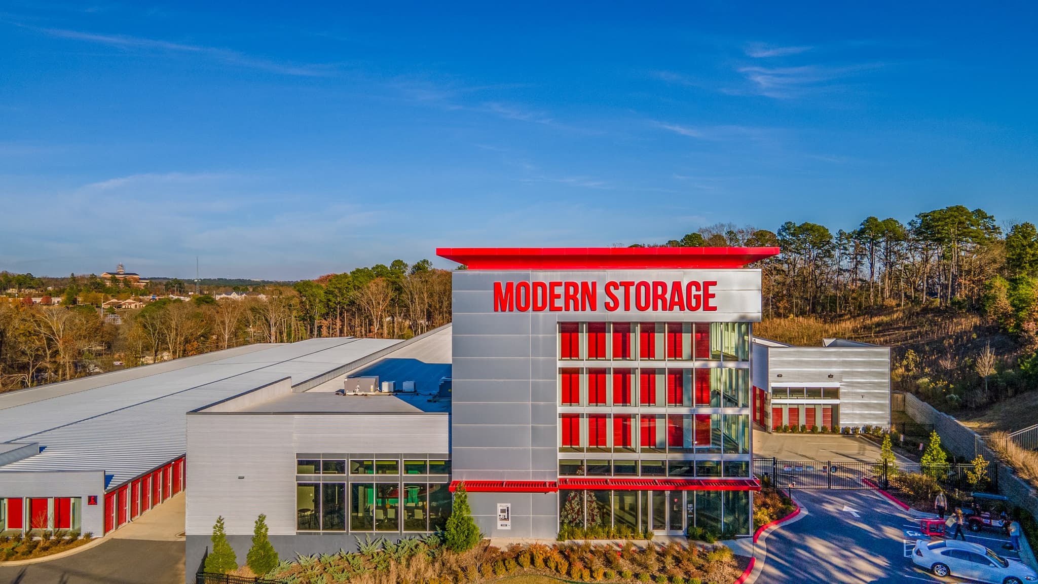 A wide-angle view of the Modern Storage® West Little Rock facility, featuring bold red branding, sleek architecture, and a forested hillside in the background under a clear blue sky.