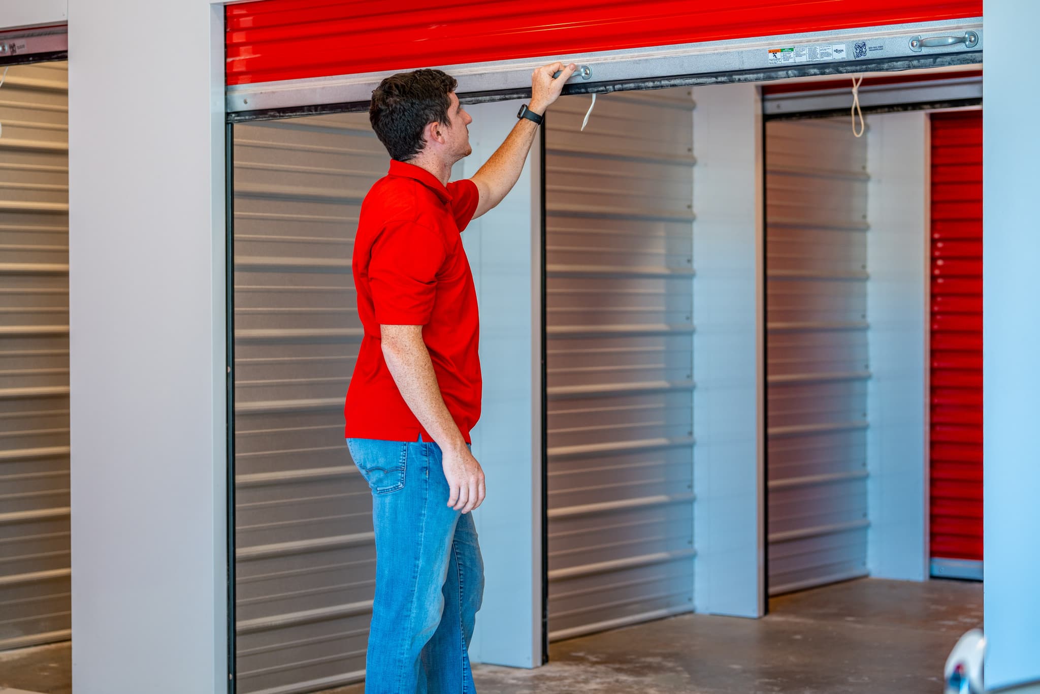Man in red shirt opening a roll-up storage unit door at a Modern Storage® facility with clean, well-lit indoor access