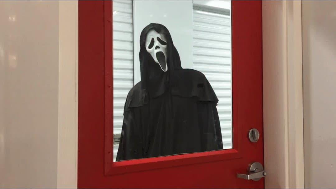 A person in a Scream mask and robe ominously peers through a red door window inside a storage facility, humorously symbolizing hidden storage fees