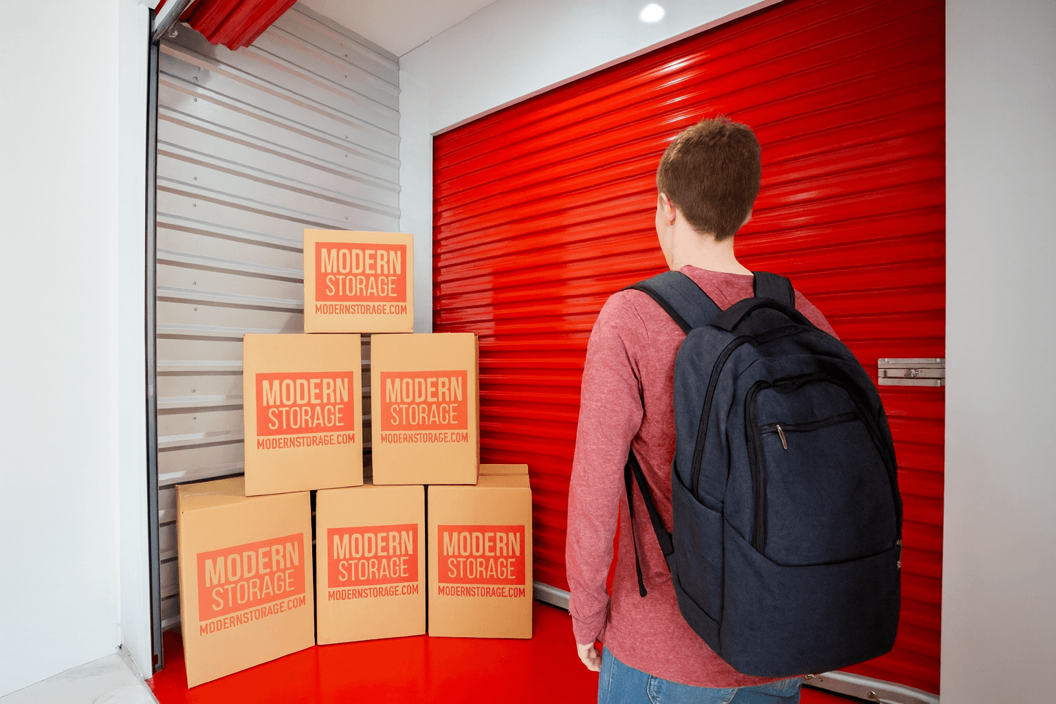 A person carrying multiple cardboard boxes into a bright red Modern Storage® unit, representing back-to-school organization a