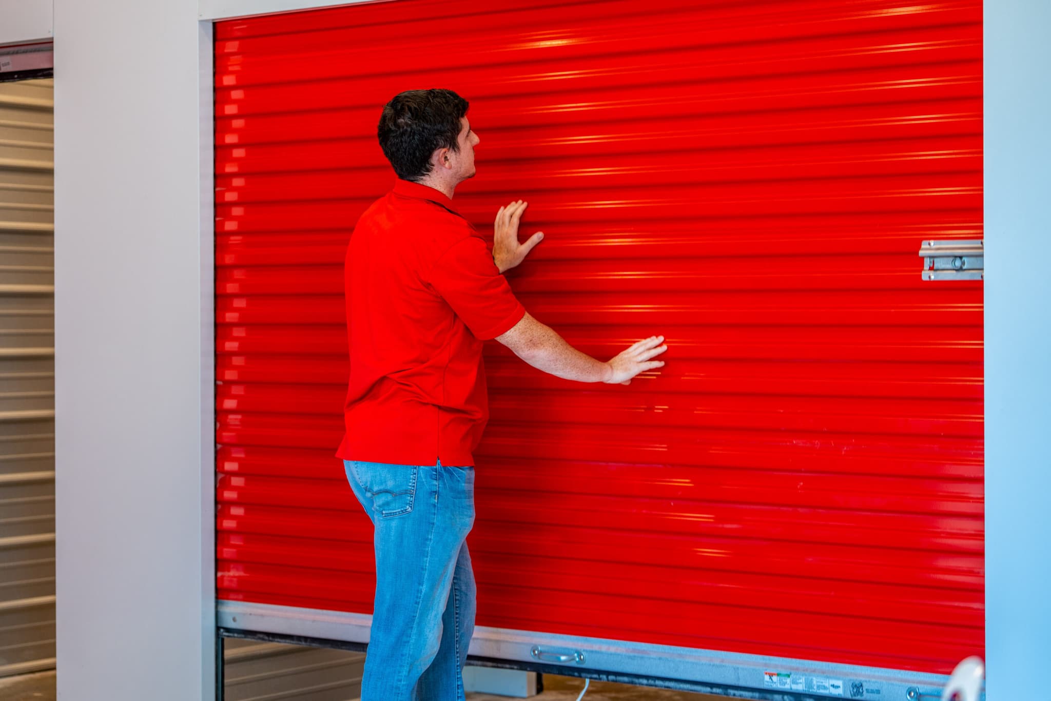 Modern Storage® employee in red polo closing a bright red roll-up storage unit door inside the facility