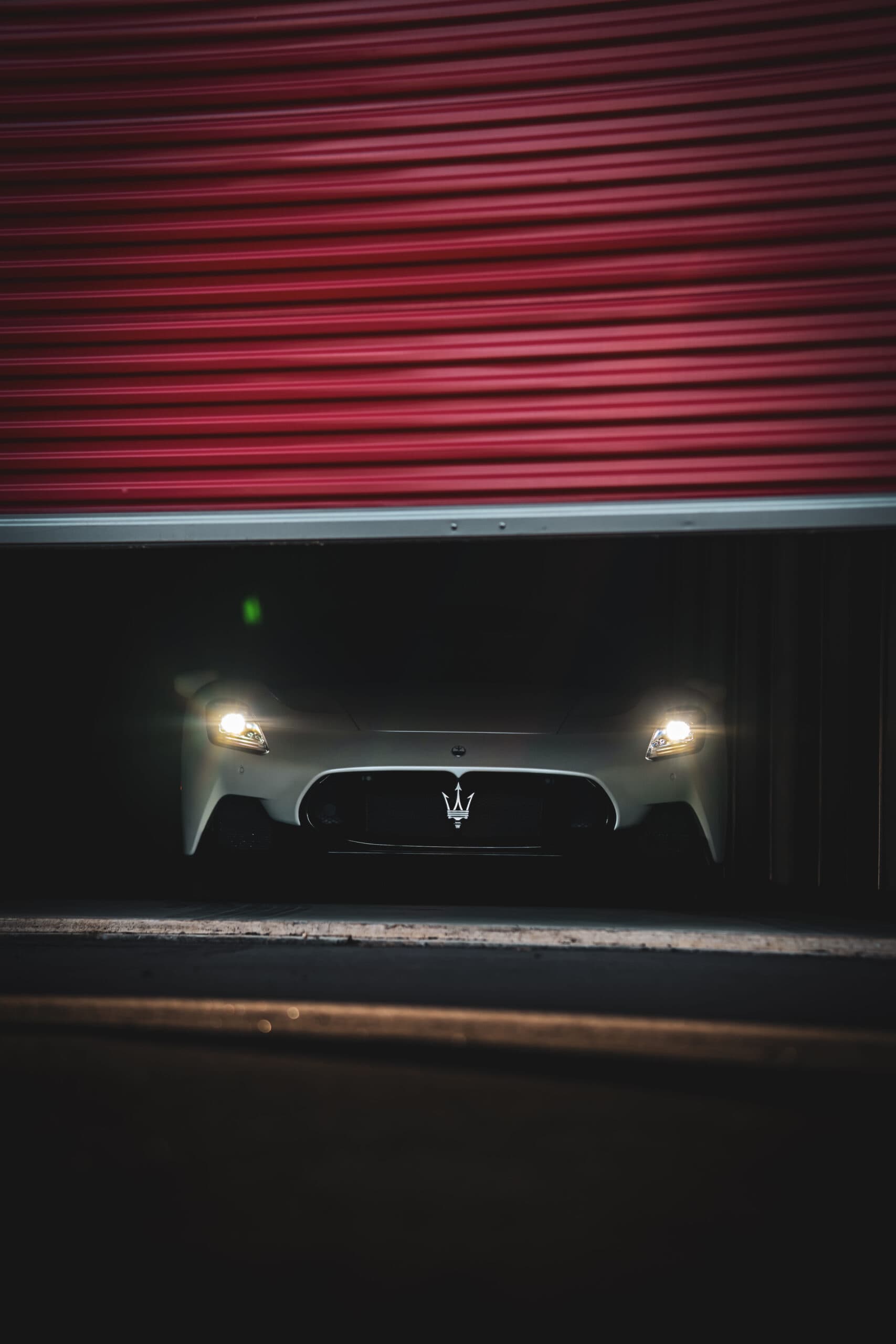 White Maserati sports car with headlights on, parked inside a secure storage unit with a red roll-up door at Modern Storage®.